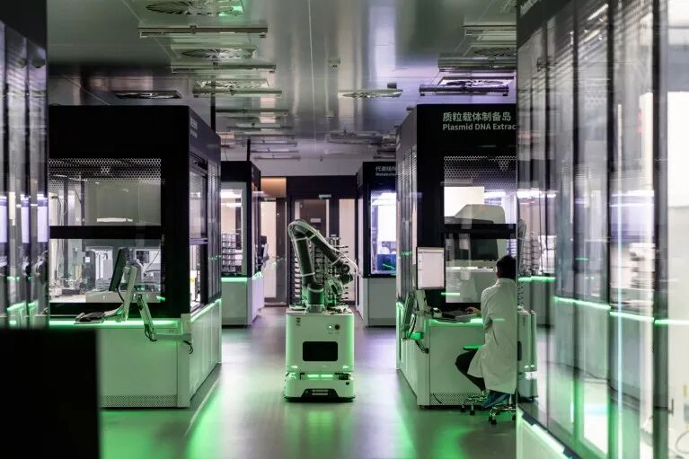 A robot is seen inside a lab, which is lit by a soft green glow, with a person in a lab coat sitting on a stool to its right