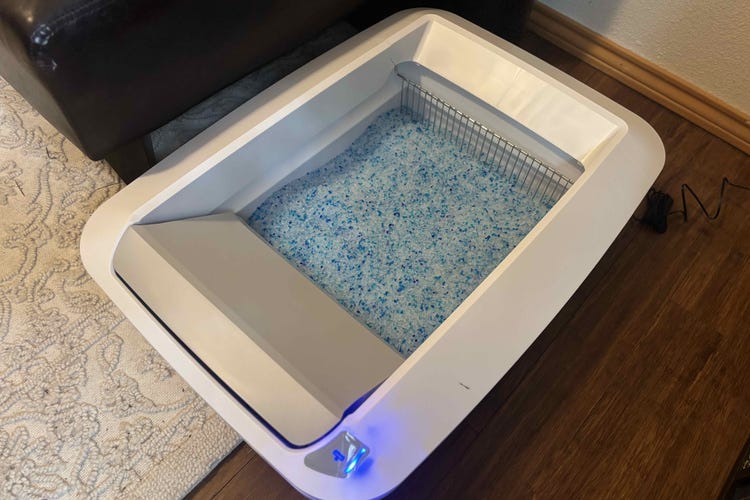 A PetSafe litterbox open and filled with blue litter on a wood floor.
