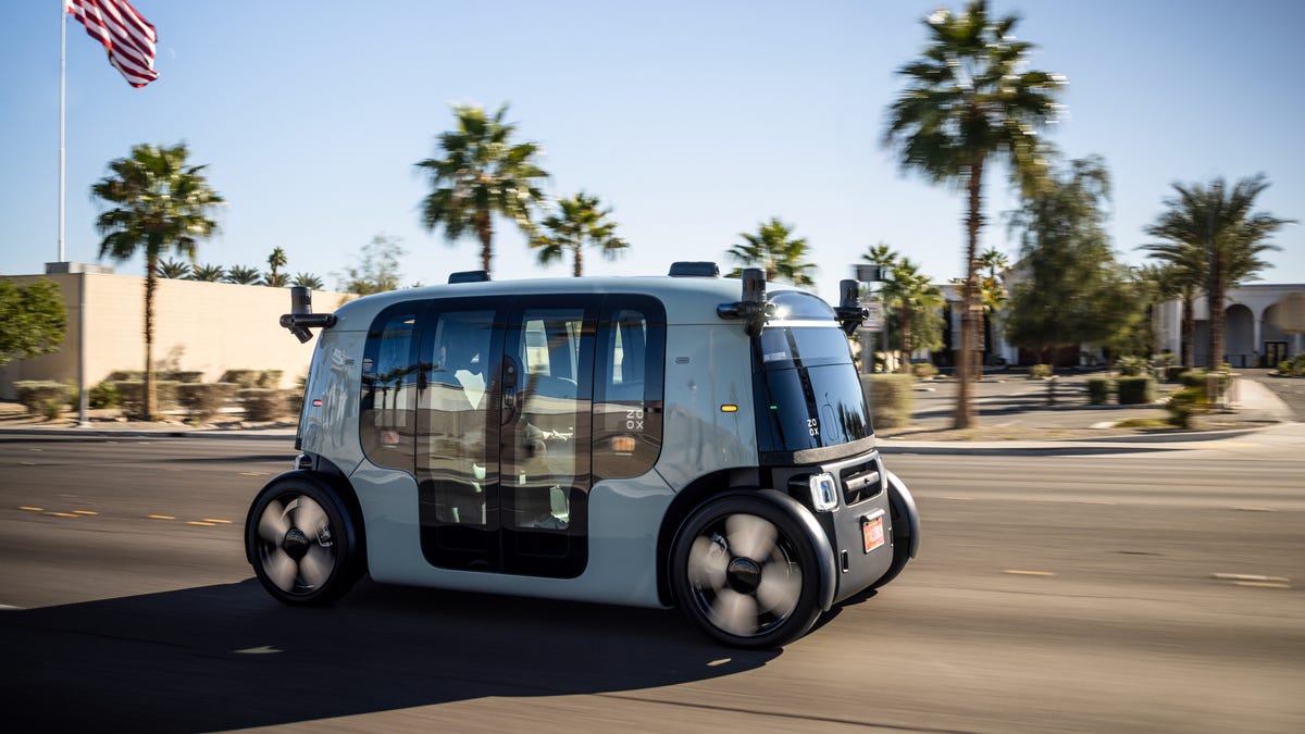 A Zoox robotaxi on the road in Las Vegas, with palm trees and an American flag in the background