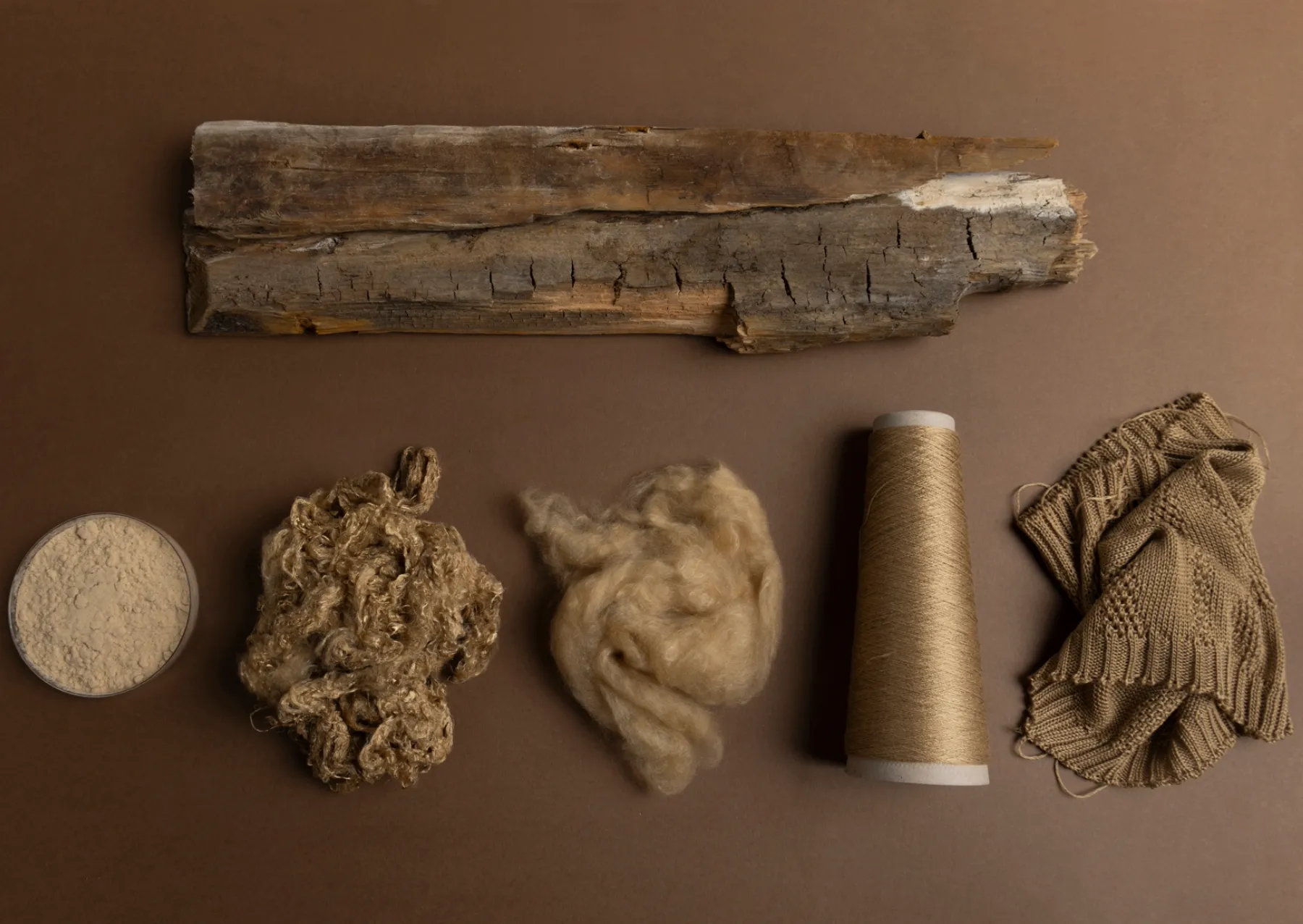 the materials used to make a dress laid out on a brown background. a piece of wood is on top, with wood pulp, fiver, yarn, and he fabric below