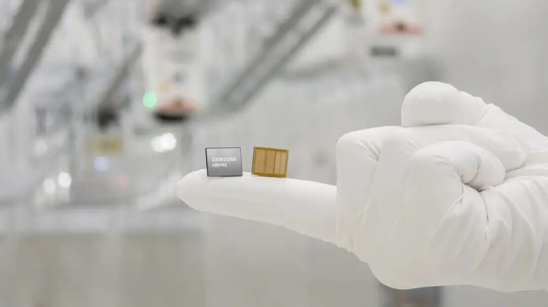 A Samsung HBM4E memory chip and a yellow circuit board placed on a gloved finger, with semiconductor manufacturing equipment blurred in the background.