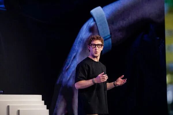 Mark Zuckerberg, wearing black-rimmed eyeglasses and a black T-shirt, gestures while speaking on a stage.